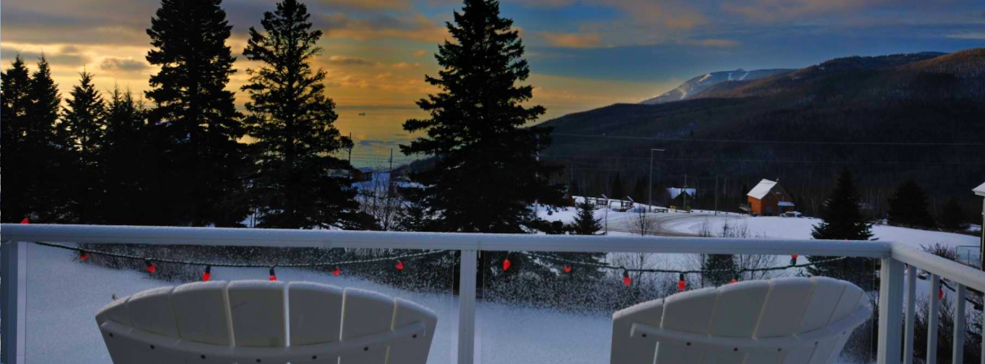 Balcon enneigé avec des chaises blanches, vue sur les montagnes et le fleuve au coucher de soleil, un environnement paisible pour un chalet à louer à Charlevoix en hiver.